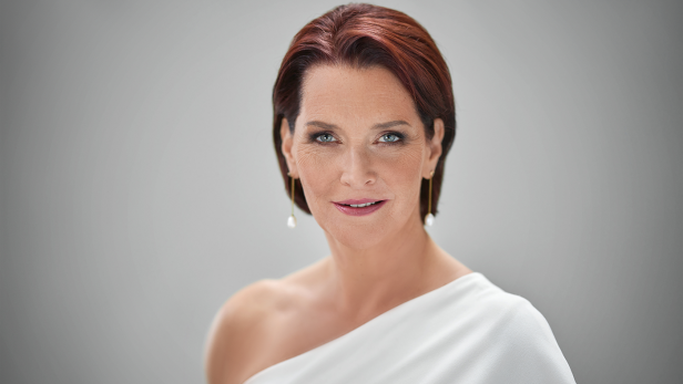 Eine Frau mit roten Haaren und einem weißen Kleid mit einem Träger lächelt in die Kamera.