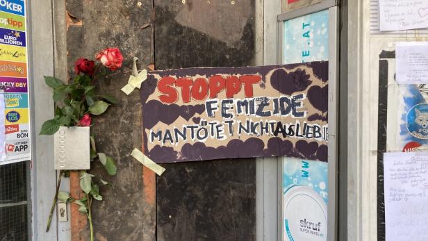 Vor der Trafik hängt ein handgemaltes Schild gegen Femizide und ein Blumenstrauß.