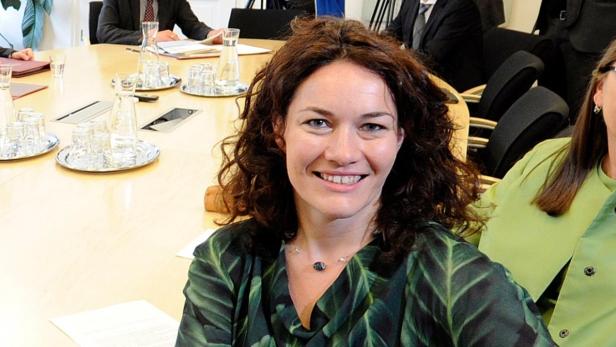 Eine Frau mit lockigem Haar lächelt in die Kamera, während sie an einem Konferenztisch sitzt.