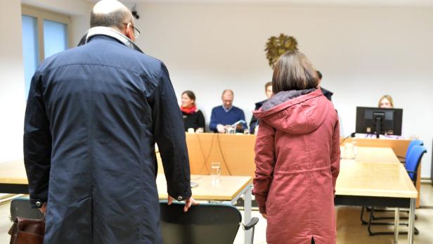 Zwei Personen stehen vor einem Tisch mit mehreren Personen im Hintergrund.