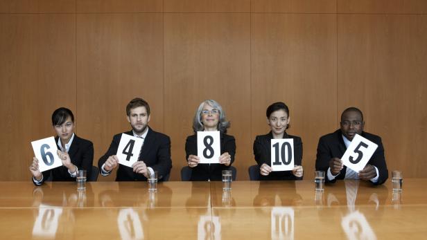 Fünf Leute in Anzügen sitzen in einem Büro und halten Schilder hoch. Die erste Frau im Anzug hält die Nummer 6, der Mann daneben die Nummer 4, die Frau in der Mitte die Nummer 8, die Frau daneben die Nummer 10 und der Mann neben ihr die Nummer 5