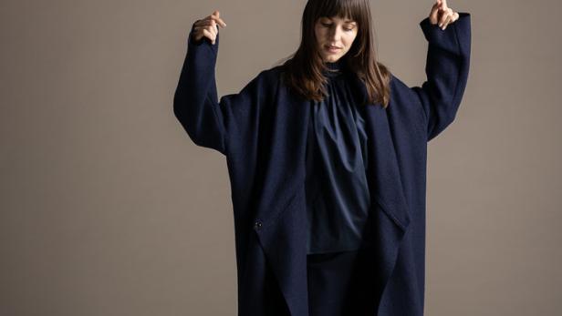 Eine Frau in einem langen, marineblauen Mantel und passender Hose posiert vor einem neutralen Hintergrund.