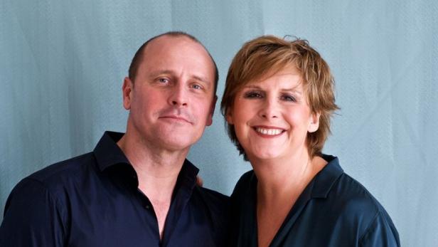 Ein Mann und eine Frau stehen lächelnd vor einem hellblauen Hintergrund.