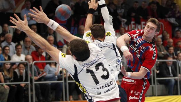 Ein Handballspieler wirft den Ball, während zwei Gegenspieler versuchen, ihn zu blocken.