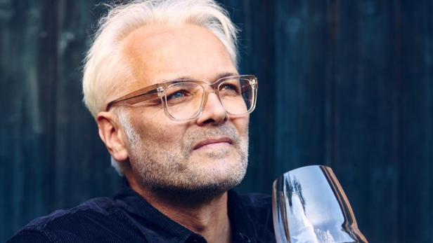 Ein Mann mit Brille betrachtet ein Glas Rotwein vor einer dunklen Holzwand.