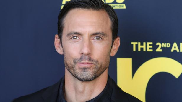 Milo Ventimiglia posiert vor einem dunkelblauen Hintergrund mit gelber Schrift.