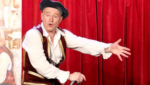 Ein Mann in historischer Kleidung gestikuliert auf einer Bühne vor einem roten Vorhang.