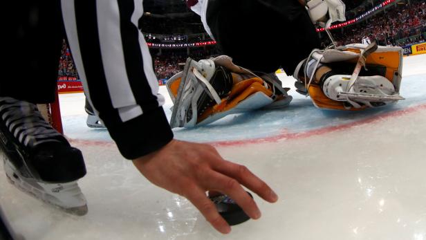 Ein Schiedsrichter legt einen Puck auf das Eis, während ein Eishockeytorwart im Hintergrund liegt.