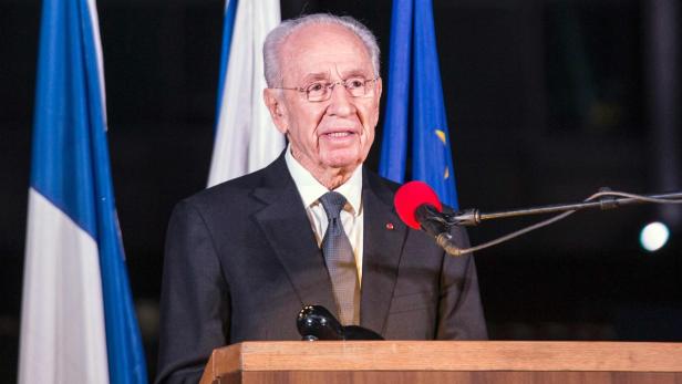 Ein älterer Mann mit Brille spricht vor Flaggen an einem Rednerpult.