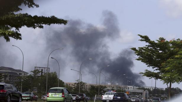Dichter, schwarzer Rauch steigt in der Ferne über einer belebten Straße auf.