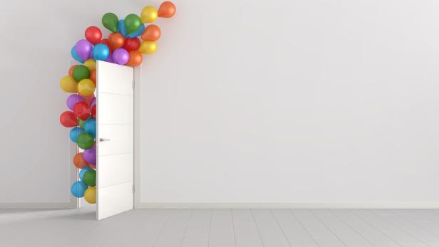 Bunte Luftballoons fiegen durch eine halb-offene weiße Tür an einer weißen Wand mit weißem Parket
