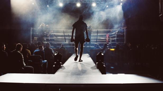 Ein Boxer auf dem Weg Richtung Boxring; links und rechts von ihm sitzt das Publikum.