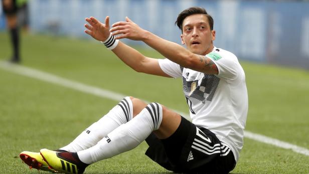 Mesut Özil sitzt auf dem Rasen und gestikuliert mit den Händen.