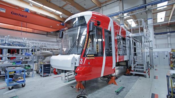 Eine rot-weiße Straßenbahn steht in einer Werkshalle auf orangefarbenen Stützen.