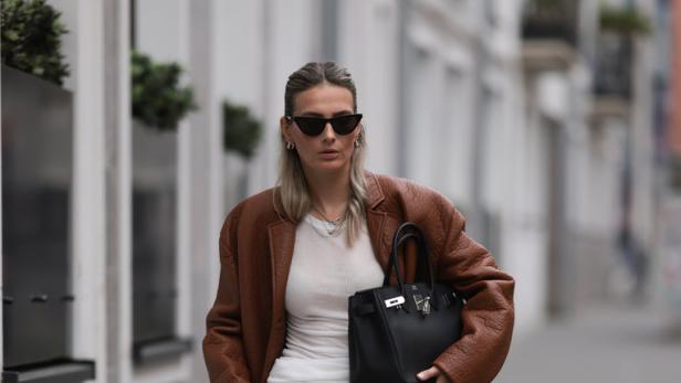 Eine Frau mit Sonnenbrille, brauner Jacke und grauer Hose geht auf dem Bürgersteig.
