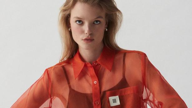 Eine Frau mit blonden Haaren trägt eine orange, transparente Bluse.