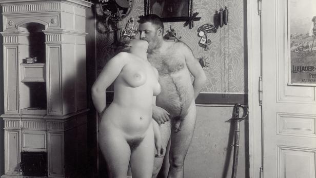 Ein nacktes Paar steht in einem Raum mit einem Kamin und einer Kuckucksuhr und küsst sich.