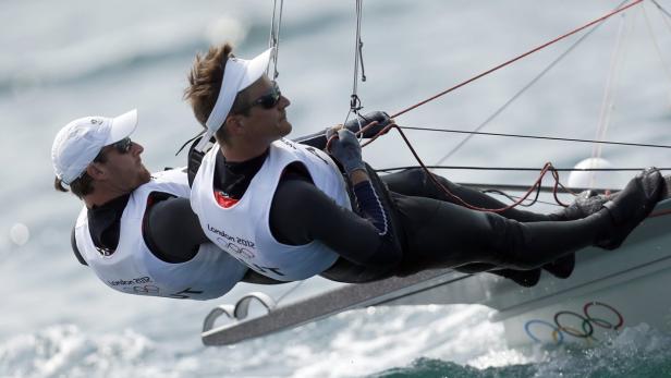 Zwei Segler in Neoprenanzügen steuern ein Segelboot mit den olympischen Ringen.