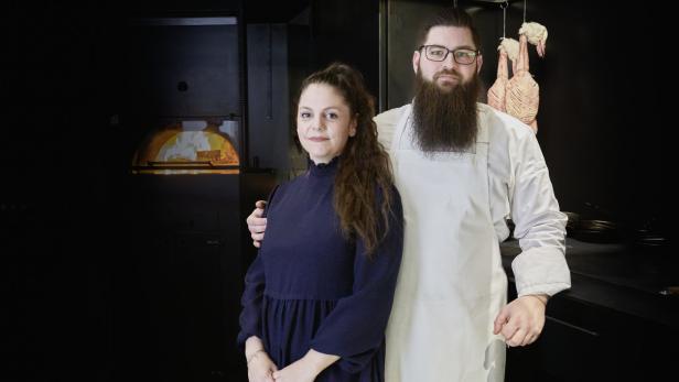 Ein Koch mit Bart und eine Frau stehen in einer dunklen Restaurantküche.