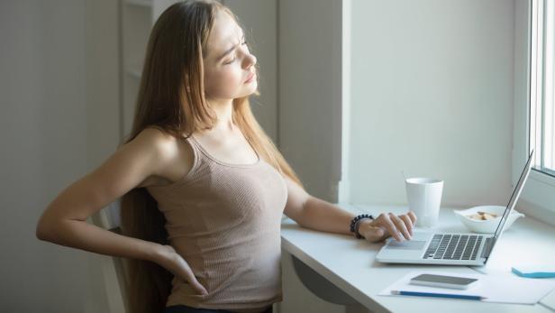Eine junge Frau sitzt vorm Computer an einem Schreibtisch und greift sich an den Rücken.