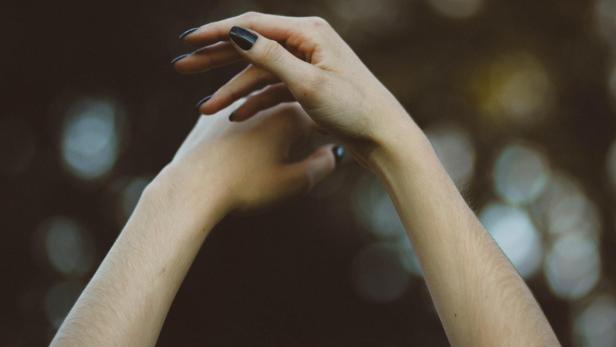 Zwei Hände mit dunklem Nagellack sind vor einem verschwommenen Hintergrund erhoben.
