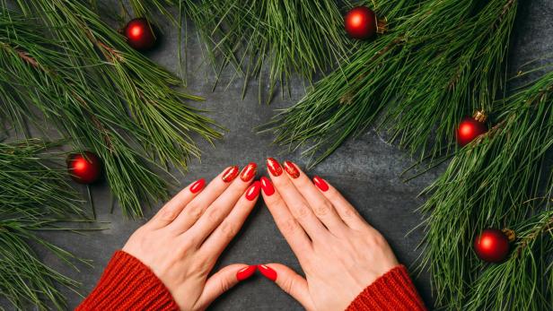 Hände mit rotem Nagellack und Weihnachtsmotiven vor Tannenzweigen und roten Kugeln.