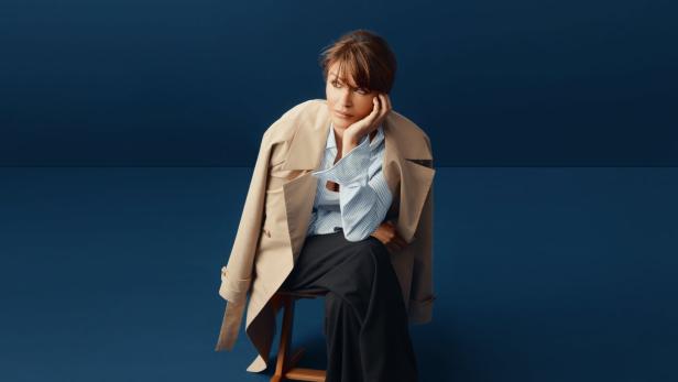 Eine Frau sitzt auf einem Stuhl, den Kopf in die Hand gestützt, und blickt nachdenklich zur Seite.