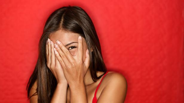 Eine junge Frau hält ihre Hände vor ihr Gesicht und schaut hervor, vor einem roten Hintergrund.