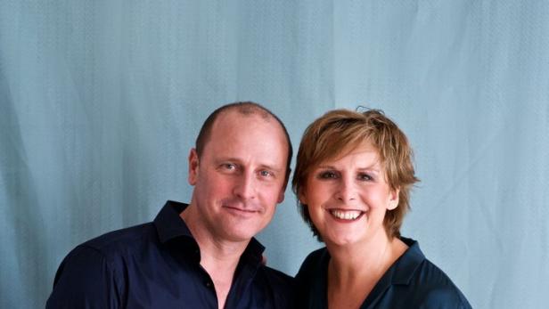 Ein Mann und eine Frau stehen lächelnd vor einem hellblauen Hintergrund.