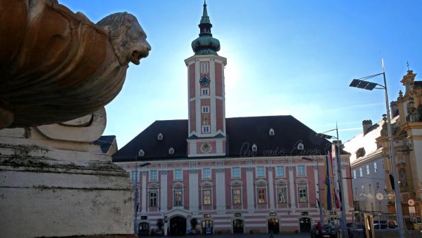 Ein rosafarbenes Gebäude mit Turm und Uhr steht hinter einem steinernen Löwenkopf im Vordergrund.