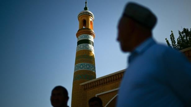 Ein verziertes Minarett steht vor blauem Himmel, im Vordergrund verschwommene Personen.