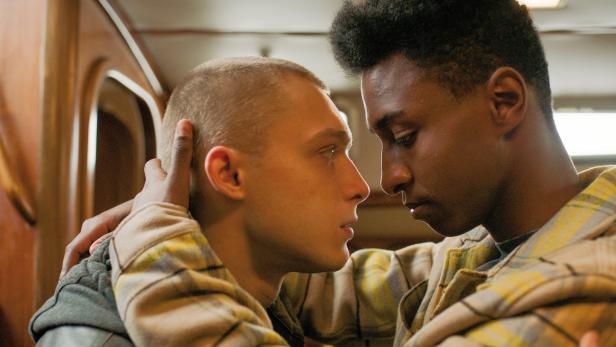 Zwei junge Männer stehen einander nah gegenüber, einer berührt den Kopf des anderen.