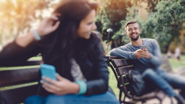 Ein lächelnder Mann sitzt mit seinem Handy auf einer Parkbank, während eine Frau neben ihm sitzt.