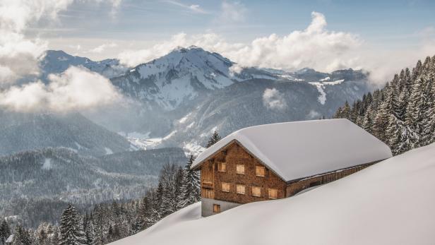 Ein schneebedecktes Holzhaus vor einer verschneiten Berglandschaft.