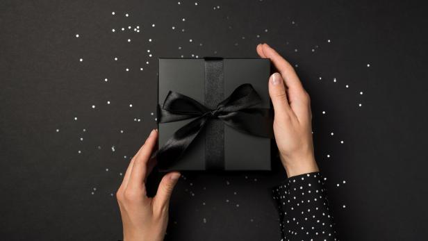 Hände halten eine schwarze Geschenkbox mit Schleife auf einem schwarzen Hintergrund mit Konfetti.