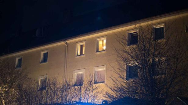 Ein Mehrfamilienhaus bei Nacht mit beleuchteten Fenstern hinter kahlen Bäumen.