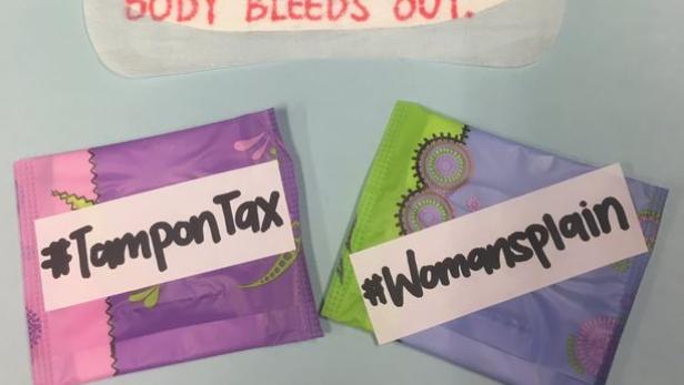 Eine Menstruationsbinde mit dem Text „Ich verbringe 5 Tage damit, mich übergeben zu wollen, während mein Körper ausblutet“ und zwei Bindenpackungen mit den Hashtags #TamponTax und #Womansplain.