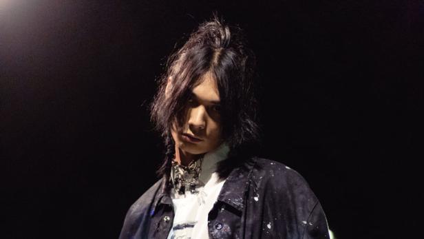 Ein junger Mann mit langen, dunklen Haaren trägt eine bemalte Jacke und ein Hemd mit Farbflecken.