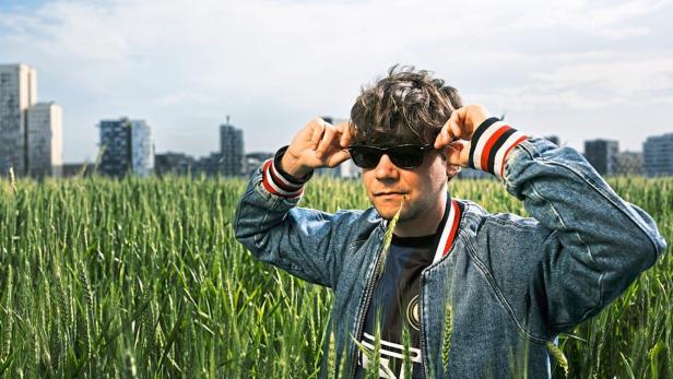 Ein Mann mit Sonnenbrille steht in einem Weizenfeld vor einer Stadtkulisse.