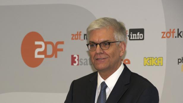 Ein Mann mit Brille und Anzug steht vor einem Hintergrund mit Logos verschiedener Fernsehsender.
