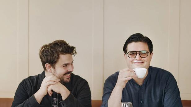 Zwei Männer sitzen an einem Tisch in einem Café, einer trinkt Kaffee.