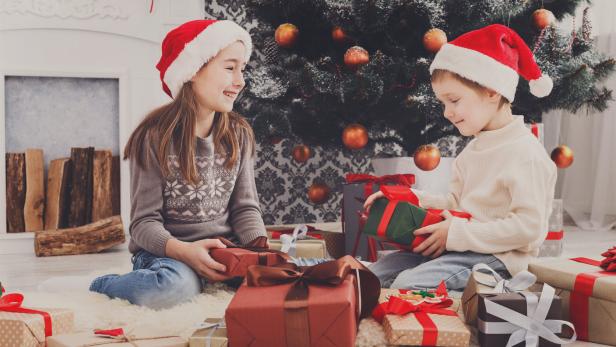 Zwei Kinder mit Weihnachtsmannmützen packen Geschenke aus.