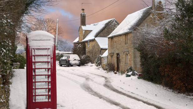 Eine rote Telefonzelle steht verschneit vor einem Dorfhaus.
