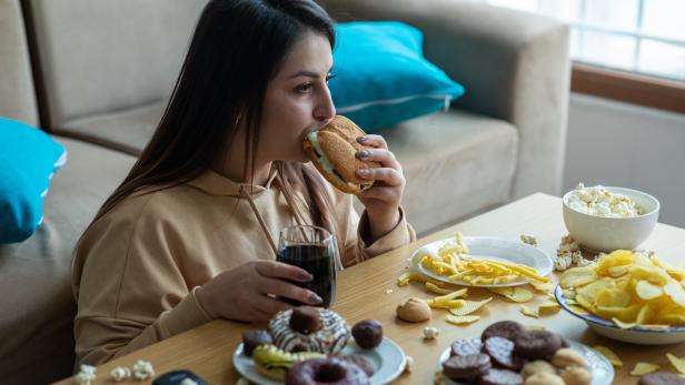Eine junge Frau sitzt vor einem Tisch mit Junk Food und isst einen Burger.