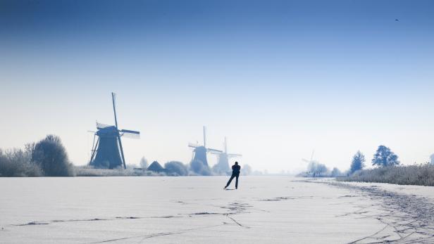 Eine Person läuft Schlittschuh auf einem zugefrorenen See mit Windmühlen im Hintergrund.
