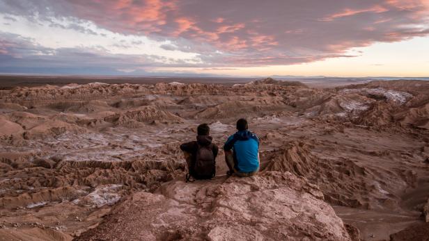 Zwei Personen sitzen auf einem Felsen und blicken auf die Wüstenlandschaft bei Sonnenuntergang.