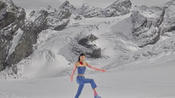 Eine Frau in Jeanskleidung watet durch den Schnee vor einer Bergkulisse.