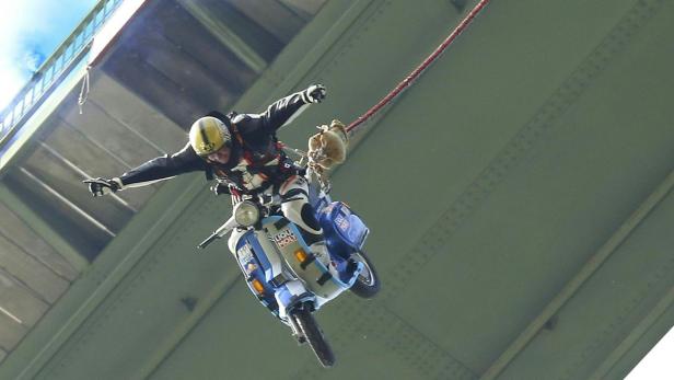 Ein Mann springt mit einem Motorroller von einer Brücke ab, gesichert durch ein Seil.