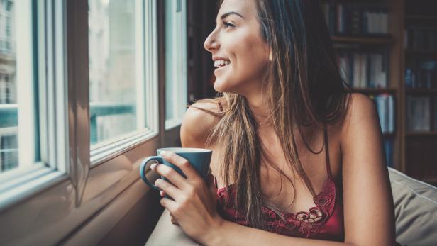 Eine Frau lächelt, während sie mit einer Tasse Kaffee am Fenster sitzt.