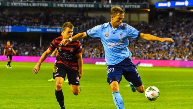 Ein Fußballspieler in einem hellblauen Trikot schießt den Ball, während ein Gegenspieler zusieht.